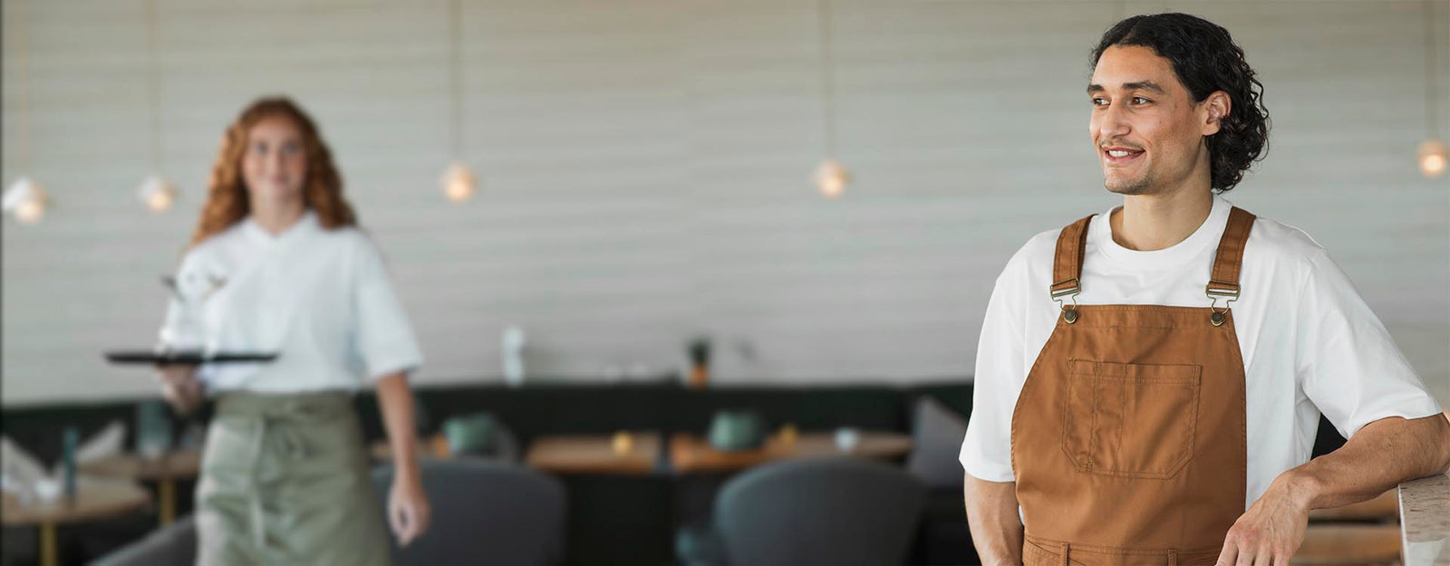 Servitör i brun förkläde ler medan han står i en modern restaurang, med en servitris som bär en bricka i bakgrunden.