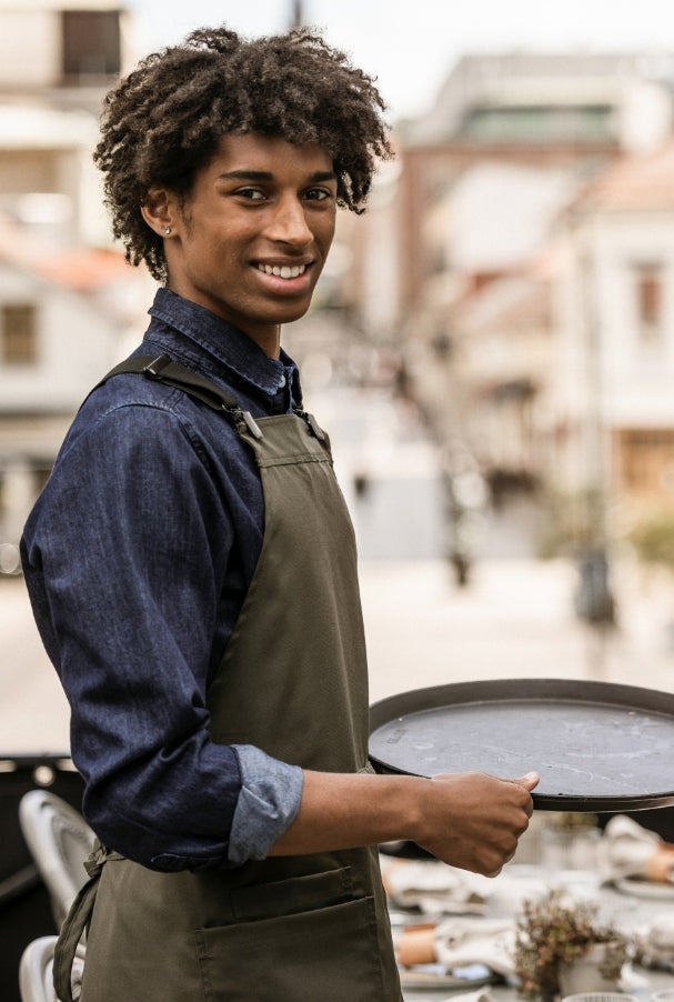 Ung servitör i olivgrönt förkläde och jeansskjorta håller en serveringsbricka utomhus på restaurangterrass.