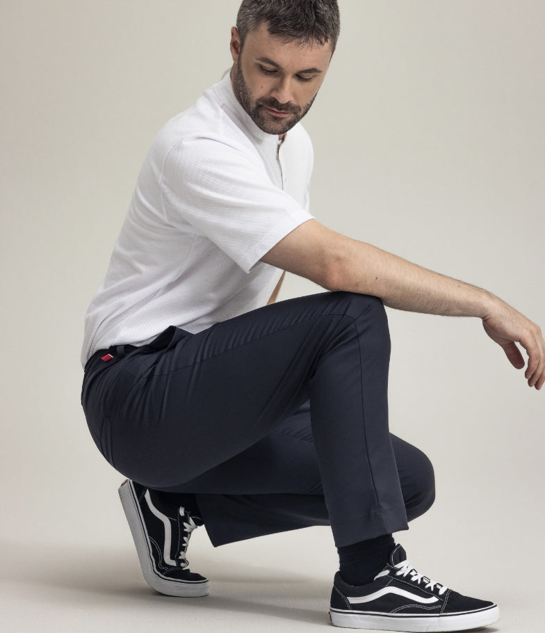 Man i vit kortärmad kockskjorta och svarta byxor sitter på huk mot en ljus bakgrund.