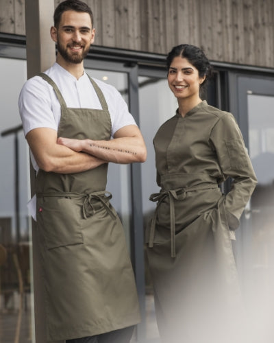 Två kockar står utanför en restaurang, klädda i matchande olivgröna förkläden och kockjackor. De ler mot kameran i ett naturligt dagsljus. Bilden uttrycker samarbete och yrkesstolthet inom restaurangbranschen.