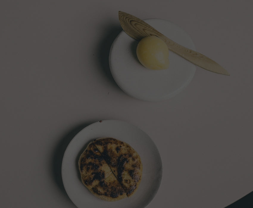 Två vita tallrikar på ett ljusrosa bord. På den ena ligger en rund, grillad brödkaka, och på den andra en gul kula smör med en träkniv ovanpå.