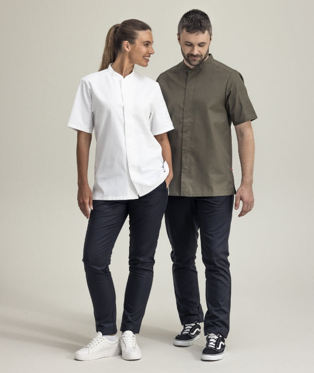 Två personer står bredvid varandra och bär arbetskläder – en i vit kortärmad skjorta och en i olivgrön. Båda har mörka byxor och sneakers, och ser avslappnade ut i en neutral studiomiljö.