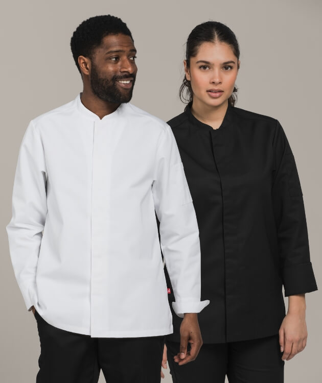 En man och en kvinna står mot neutral bakgrund, iklädda klassiska kockjackor – den ena i vitt och den andra i svart. De ser självsäkra ut och visar upp stilrena, unisex-designade plagg för kökspersonal.